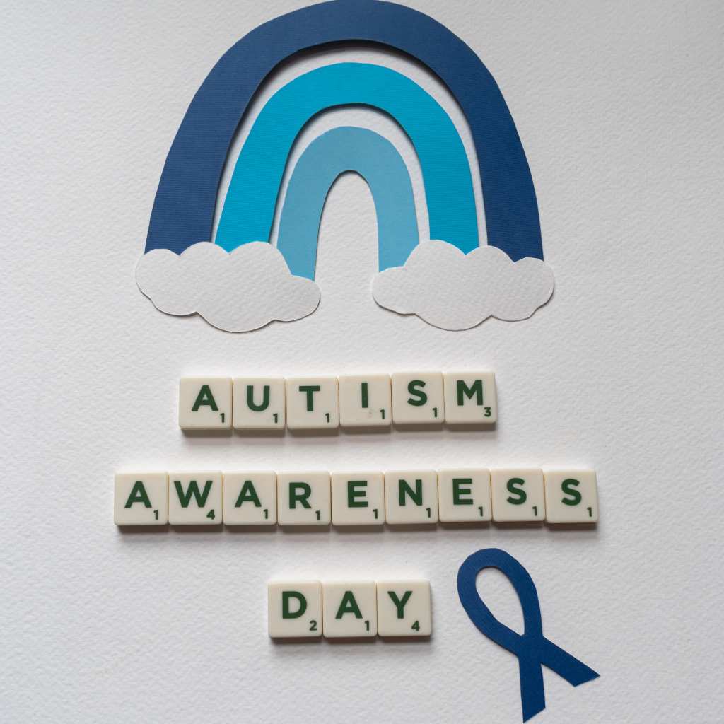 Blue paper rainbow and clouds with letter tiles reading Autism Awareness Day beside a blue ribbon